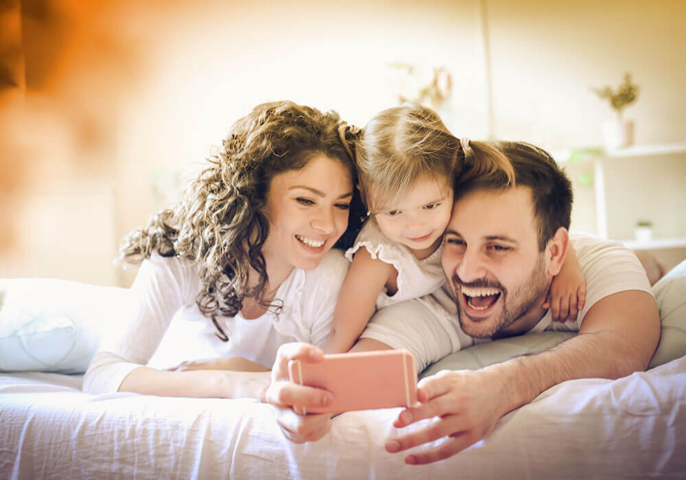 Image of a husband, wife, and young daughter all lying in bed and smiling at something they are seeing on a smartphone.
