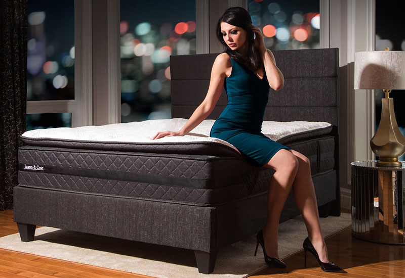 Image of a female sitting on the edge of the Logan & Cove luxury hybrid pillow-top mattress with a colourful city night sky in the background.